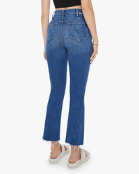 Someone wears Mother’s The Hustler Ankle Fray in Grasping At Straws—high-rise flare jeans with raw, frayed hems—paired with white sandals and a black top, seen from the back against a plain background.