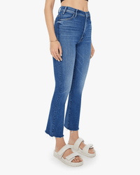 A person wearing The Hustler Ankle Fray in Grasping At Straws by Mother, featuring frayed hems and paired with white sandals, stands against a plain white background. Only the lower body is fully visible.