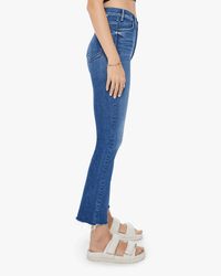 A person stands in profile wearing Mother’s The Hustler Ankle Fray in Grasping At Straws high-rise flare jeans with a raw hem, paired with a black top, bracelet, and white sandals against a plain white background.