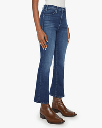 A person wears Mother’s The Hustler Ankle in Heirloom jeans, a white top, and brown leather ankle boots, standing against a plain white background with their left hand by their side.