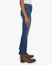 A person is standing sideways against a plain white background, wearing Mother’s The Hustler Ankle in Heirloom jeans, a white top, and brown leather ankle boots, showing the left profile of their body.