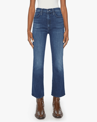A person wearing The Hustler Ankle in Heirloom jeans by Mother, brown leather boots, and a white top stands with arms relaxed by their sides against a white background.