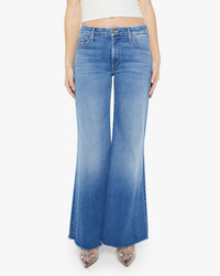 A person wears Mother’s The Lil Roller Fray in Work Hard Play Hard—high-waisted, light blue stretch denim jeans with a frayed hem—paired with a white crop top and pointed-toe shoes, shown waist to feet on a white background.