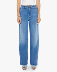 Someone stands against a plain white background wearing The Major Prep Zip Sneak in Ready Set Go jeans by Mother, featuring a high-waisted, wide straight-leg blue denim design with front and side pockets, paired with a white top and open-toe shoes.