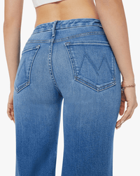 A woman seen from the back wears MOTHER's The Roller Fray in Work Hard Play Hard—high-waisted blue wide-leg jeans with a raw hem, stitched back pockets, and a subtle fade. She pairs them with a white crop top and a bracelet on her right wrist.