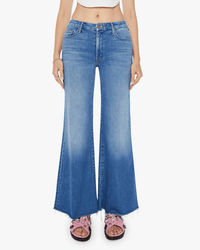 Someone is wearing Mother’s The Roller Fray in Work Hard Play Hard jeans—high-waisted, light blue wide leg denim with a raw hem—paired with pink sandals featuring black soles and flower accents, against a white background.