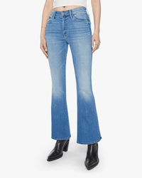 A person wearing The Weekender in Layover jeans by Mother, a white cropped top, and black heeled ankle boots stands against a plain white background.
