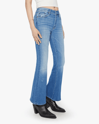 A person wearing Mother’s The Weekender in Layover jeans, a white sleeveless top, and black heeled ankle boots stands against a plain white background.