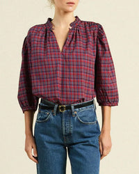 A person wears the Trovata Birds of Paradis Orly Blouse in Mulled Wine Plaid, featuring a ruffled collar and three-quarter sleeves, tucked into high-waisted blue jeans with a black belt.