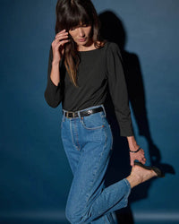 A woman with long brown hair wears the Nation LTD Amelie Envelope Neck Top in Jet Black, paired with high-waisted blue jeans. She stands against a dark blue background, looking down as she adjusts her black high-heeled shoe.