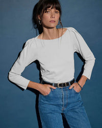 A woman stands against a blue background wearing the Nation LTD Amelie Envelope Neck Top in White, blue high-waisted jeans, a black belt, and hoop earrings, with her hands in her pockets.