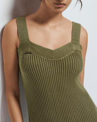 A woman models the Caresse Sweater Dress in Capers by Nation LTD, a sleeveless ribbed olive green knit dress with wide straps and a sweetheart neckline, standing against a white wall. Her head is partially cropped from the image.