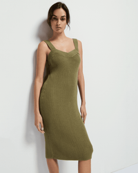 A woman stands against a white wall, confidently wearing the Nation LTD Caresse Sweater Dress in Capers—a ribbed, sleeveless, Pima cotton dress with wide straps and a fitted silhouette—looking at the camera.