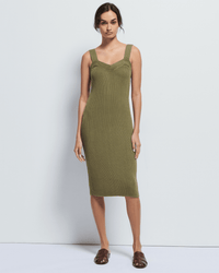 A woman stands against a plain background wearing the Caresse Sweater Dress in Capers by Nation LTD, a sleeveless, olive green ribbed knit Pima cotton dress, paired with brown open-toed flats. She faces forward with arms relaxed at her sides.