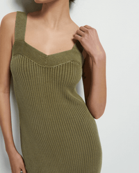 A woman models the Nation LTD Caresse Sweater Dress in Capers—a sleeveless, ribbed, olive-green Pima cotton style—standing against a white background with one hand on her shoulder and her face partly out of frame.