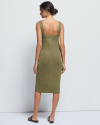 A woman with her hair in a low bun stands with her back to the camera, wearing the Nation LTD Caresse Sweater Dress in Capers and black sandals, set against a plain light background.