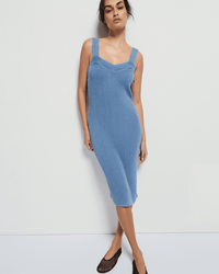 A woman stands against a white background wearing the Nation LTD Caresse Sweater Dress in Faded Denim, a sleeveless ribbed Pima cotton midi dress with thick straps, paired with black flats. She tilts her head, arms relaxed at her sides.