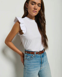 A woman with long brown hair wears the Nation LTD Paulette Tank New Perfect Slub in White, tucked into high-waisted light blue jeans with a brown belt, standing against a plain light-colored wall.