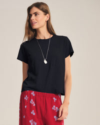 A woman with light brown hair wears the Velvet by Graham & Spencer Marika Short Sleeve Crew Neck Crop Top in black, paired with red floral pants and a pendant necklace, standing against a plain background.