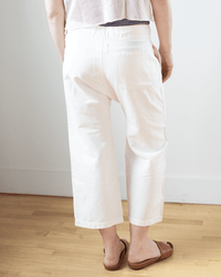 A person stands indoors on a wooden floor, wearing NSF Akari Tie Waist Pants in Ivory and brown slip-on sandals. The photo is taken from behind, focusing on the lower half of the body.