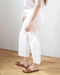 A person stands on a light wooden floor against a white wall, shown from the shoulders down, wearing NSF's Akari Tie Waist Pants in Ivory with a white mesh top and brown sandals.
