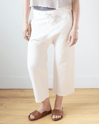 A person stands on a wooden floor wearing NSF Akari Tie Waist Pants in Ivory with a drop crotch and drawstring waist, a cropped top, and brown open-toe sandals. Their face is not shown in the image.