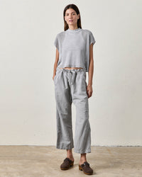 A woman with straight brown hair wears NSF's Akari Tie Waist Pants in Pale Grey, featuring a cropped inseam and loose fit, paired with a textured gray short-sleeve top and brown shoes, standing against a plain light background with hands in her pockets.