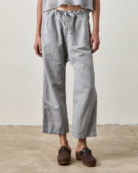 A person stands indoors on concrete wearing NSF's Akari Tie Waist Pants in Pale Grey, styled high-waisted with a cropped inseam. Their look is completed with a cropped top and dark brown clogs, face not visible.