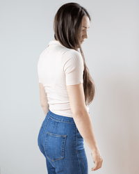 A woman with long brown hair wears the NSF Alden S/S Fitted Snap Polo in Pearl and high-waisted blue jeans, standing slightly turned against a plain light background.