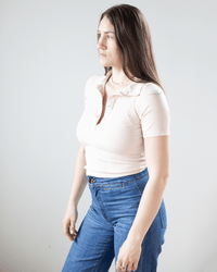 A woman with long brown hair wears the NSF Alden S/S Fitted Snap Polo in Pearl and blue jeans, standing against a plain light background while looking slightly to the side.