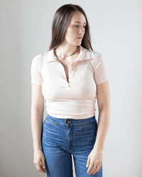 A woman with long brown hair wears the NSF Alden S/S Fitted Snap Polo in Pearl and blue jeans, standing against a plain white background. She looks slightly to her left with a neutral expression.