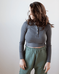 A woman with wavy brown hair, wearing the NSF Ali Fitted L/S Rib Polo in Faded Black and high-waisted green pants, stands against a light background, looking to her left.
