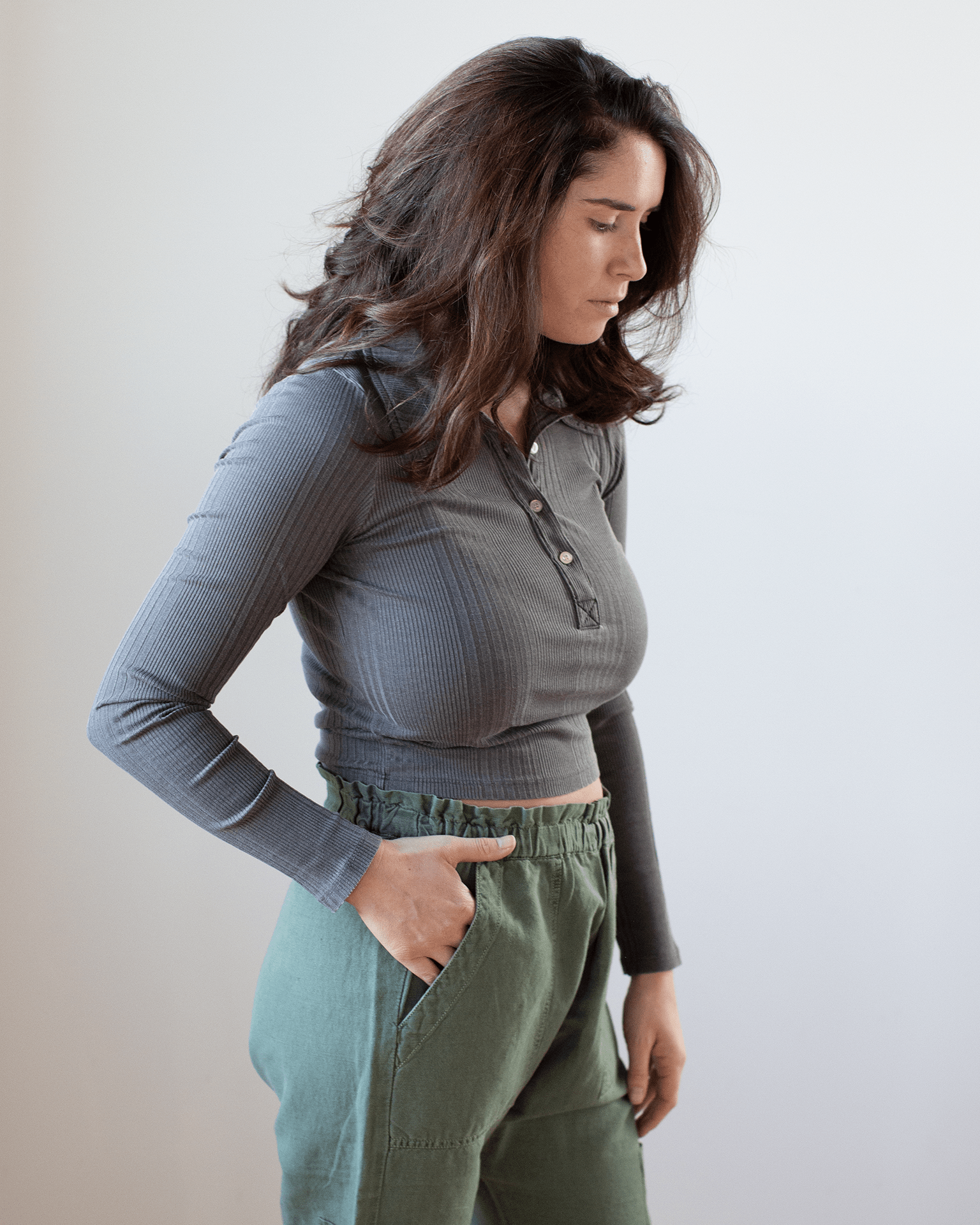 A woman with wavy brown hair wears the NSF Ali Fitted L/S Rib Polo in Faded Black and green pants, standing sideways with one hand in her pocket against a plain, light background.