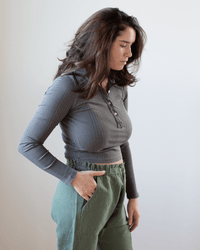 A woman with wavy brown hair wears the NSF Ali Fitted L/S Rib Polo in Faded Black and green pants, standing sideways with one hand in her pocket against a plain, light background.