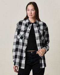 A woman models the NSF Andres Plaid Shirt in Vette, layered over a black top and black pants. She stands against a plain light background, gazing to the side with one hand adjusting her shirt.