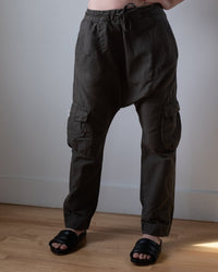 A person wearing NSF Eddy Pants in Mill Green with utility pockets and black slide sandals stands on a wooden floor against a plain, light-colored wall.