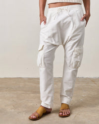 A person wearing NSF’s Eddy Pants in Soft White stands on a concrete floor against an off-white wall, styled with tan sandals. Only the lower body appears, featuring the pants’ loose fit and utility pockets.