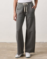 Wearing NSF’s Franky Drawstring Baker Sweats in Faded Olive—wide leg with an elastic waistband, front patch pockets, and a white drawstring—paired with a white tank top and sneakers, someone stands on a concrete floor against a light wall.