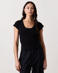 A woman with long dark hair wears the NSF Gia Fitted Rib U Neck in Black and black pants, standing against a plain light-colored wall and looking directly at the camera with a neutral expression.