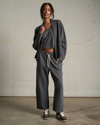 A woman stands indoors against a plain wall, wearing a loose dark gray lounge set with wide-leg pants, a matching top, and the perfectwhitetee Olivia Fleece Short Cardigan in Charcoal. She pairs the outfit with brown sneakers and has her hair tied back.