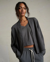 A woman with wavy hair models the perfectwhitetee Olivia Fleece Short Cardigan in Charcoal over a gray V-neck crop top and drawstring pants, posing with hands in pockets against a plain light background.