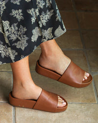 A person wearing a black skirt with white floral patterns and chic beek.. Pelican in Tan slides stands on tan tiled flooring.