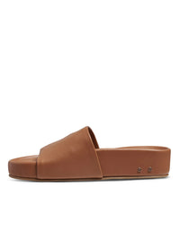The Pelican in Tan by beek. is a brown leather slide sandal with a wide, seamless upper strap, open toe, and two small metal rivets near the heel, shown in side profile on a white background.