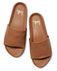 A pair of the Pelican in Tan slides by beek.—tan, open-toe with wide leather straps and flat soles—are shown from above on a white background. The insoles display beek. in gold script, offering all-day comfort and effortless style.