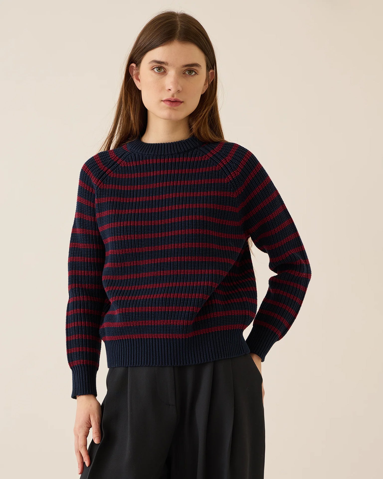 A woman with long brown hair wears the Demylee Phoebe Stripe Cotton Sweater in Navy/Burgundy and black pants, standing against a plain light background. She faces forward with a neutral expression.