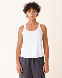 A person with short curly hair stands facing forward against a plain light background, wearing Prairie Underground’s Layering Racerback Tank in White and dark pants.
