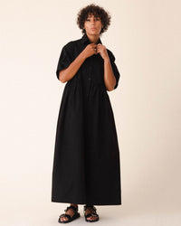 A person with curly hair wears the Prairie Underground Que Sera Sera Dress in 12am—an oversized, loose-fitting black maxi shirtdress with short sleeves and buttons—paired with black sandals, standing against a neutral background.