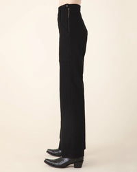 A person in profile wears Prairie Underground's Yr Arrow Pant in Black—a high-waisted style with a side zipper—paired with black pointed-toe boots, set against a plain light background.