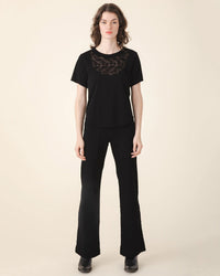 A person with wavy brown hair stands against a beige background, wearing a lace-trimmed black top and Prairie Underground's Yr Arrow Pant in Black high-rise flared jeans, paired with black shoes.