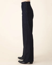 A person stands sideways wearing Prairie Underground's Yr Arrow Pant in Denim and black pointed-toe boots against a plain light background.